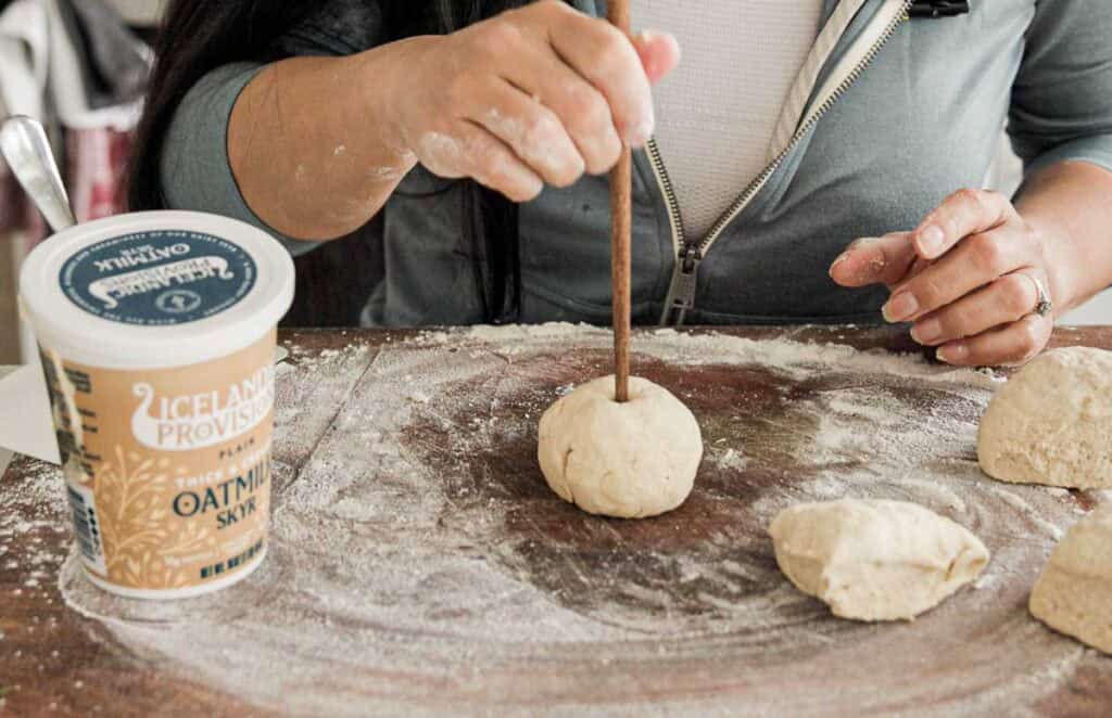 A person is shaping dough balls on a floured surface. They use a wooden stick to make an indentation in the dough. A container of Icelandic Provisions Oatmilk Skyr is visible on the left.